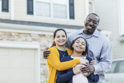 An 11 year old mixed race Hispanic and African-American girl standing with her parents outside their home. Mom is a mature Hispanic woman and dad is a mature African-American man. They are both in their 40s, happy and proud of their daughter.  All are smiling at the camera.